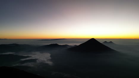 Drone-view-in-Guatemala-flying-over-a-volcano-crater-volcano-mountains-in-the-horizon-at-sunrise-covered-by-mist