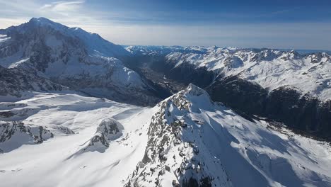 An-expansive-aerial-view-over-a-stunning-alpine-landscape,-showcasing-a-sharp-mountain-ridge-leading-to-snow-laden-peaks-under-a-clear-sky