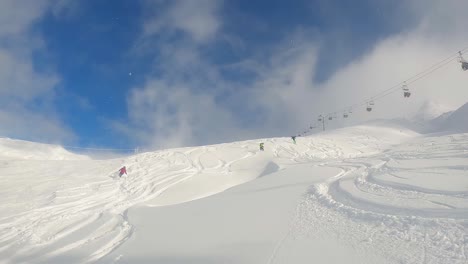 Group-of-skiers-descending-powder-off-piste,-leaving-tracks,-with-chairlift-in-background-on-clear-sunny-day