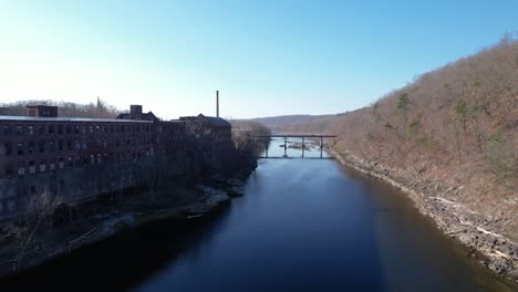 Flying-along-river-view-of-millhouses-and-branching-trees-with-rocks-on-riverbanks-and-a-bridge-crossing-next-to-a-chimney-in-the-background