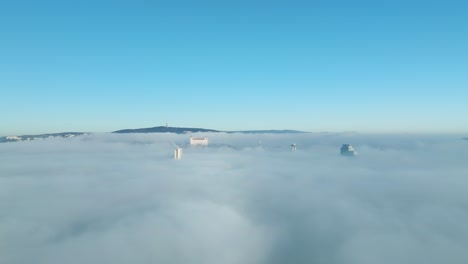Sunlit-aerial-journey:-Drone-moves-forward-towards-the-Bratislava-castle-and-buildings-peeking-through-thick-inversion-clouds,-emphasizing-the-cinematic-beauty-of-urban-exploration