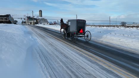Toma-Aérea-De-Seguimiento-De-Un-Carruaje-Tirado-Por-Caballos-En-El-País-Amish-Con-Nieve-En-Invierno.