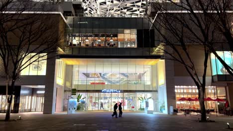 Vista-Nocturna-De-La-Fachada-De-Un-Edificio-Moderno-E-Iluminado-Con-Gente-Caminando,-En-Yokohama,-Japón