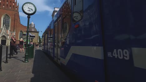 Blue-trolly-passes-an-old-railway-clock-by-a-busy-street-in-Krakow,-Poland