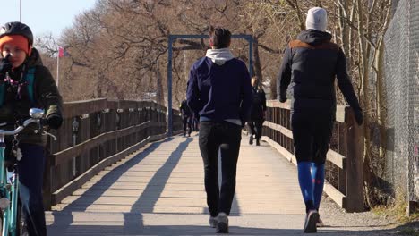 Father-and-son-ride-bikes,-others-walk,-on-wooden-bridge-by-water-in-Stockholm