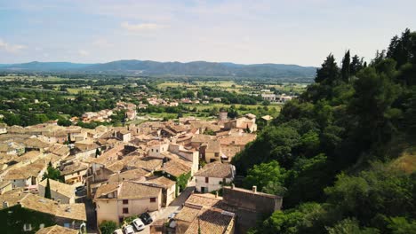 Sweeping-panoramic-view-of-tan-grey-roofs-along-forested-hillside-in-Cadenet-Provence-France,-aerial