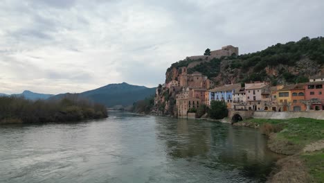 River-flowing-by-the-historic-Miravet-town-in-Tarragona,-Spain,-with-mountains-in-the-backdrop,-overcast-sky