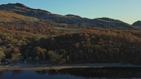 Aerial-Panning-Shot-Along-the-Shores-of-Lake-Lomond-and-The-Trossachs-National-Park-with-Autumn-Forest-in-the-Warm-Sunlight,-Scotland