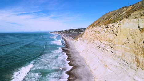 Drone-descends-along-heavily-eroded-sea-cliffs-as-calm-ocean-waves-crash-up-against-grey-sandy-beach-in-California