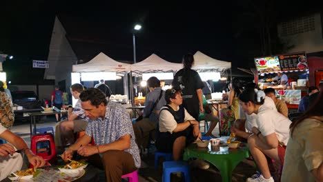 Extranjeros-Y-Locales-Comiendo-En-Asientos-Bajos-En-El-Típico-Mercado-Nocturno-De-Tailandia