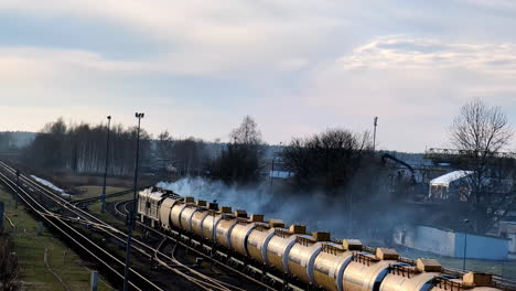 Old-Vapor-Train-arrives-slow-to-a-countryside-town-in-autumn-landscape-dry-trees-skyline,-railways-in-daylight-morning-cold-atmosphere-aerial-drone-view