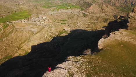 Un-Hombre-Sentado-Peligrosamente-En-El-Borde-De-Un-Promontorio-De-Roca-Del-Abismo-Del-Acantilado-Con-La-Vista-Del-Campo-De-La-Aldea-Rural-En-El-Paisaje-Natural-De-Las-Tierras-Altas-De-Irán,-Una-Aventura-En-La-Naturaleza,-Senderismo-Aéreo