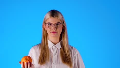 Smart-looking-blonde-caucasian-young-woman-with-glasses-plays-with-an-orange-happy,-infinite-background-chroma-blue-portrait-shot