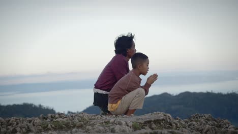 Filipino-child-claps-hand-squatting-next-to-parent-as-they-look-out-across-mountains,-blurred-background