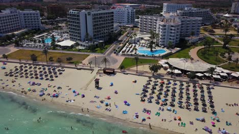 Cala-major-beach-with-sunbathers,-umbrellas,-clear-skies,-and-resort-backdrop,-aerial-view