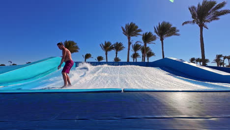 Man-surfing-on-artificial-wave-pool-with-palm-trees-in-background,-sunny-day,-resort-vibes