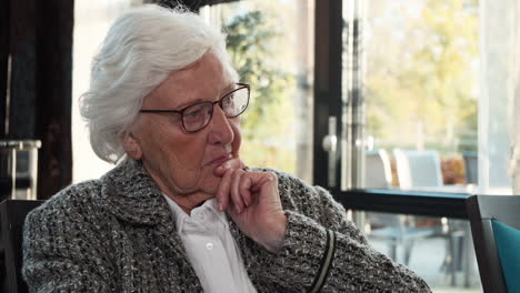 Aged-woman-sitting-in-living-room-of-nursing-home-holds-chin-and-looks-concerned