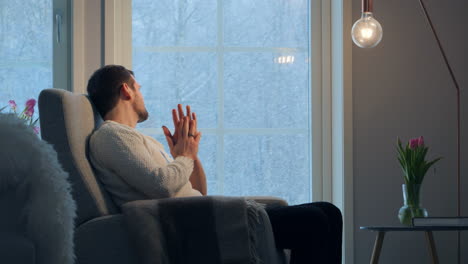 Man-in-sweater-contemplating-by-window-with-heavy-snow-outside,-serene-indoor-scene,-daylight