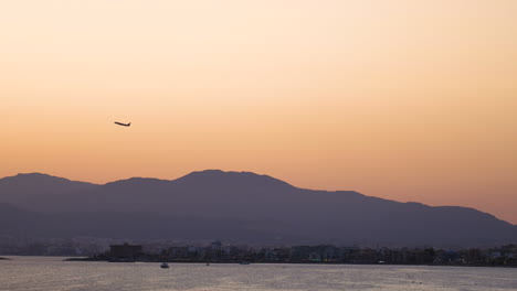 Silhouetted-airplane-taking-off-over-hazy-Mallorca-mountain-range-across-golden-orange-skyline