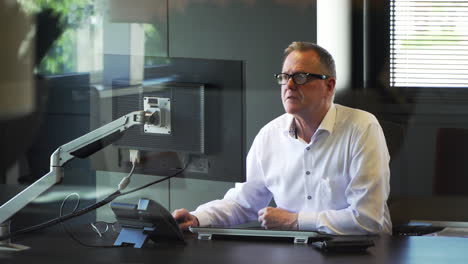 Director-sits-in-his-own-glass-office-looking-at-computer-screen-reading-email