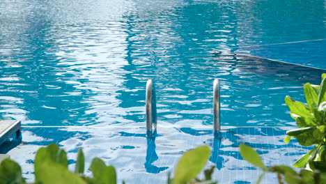 Close-up-of-Swimming-Pool-Chrome-Steel-Handrail-Descending-Into-Tortoise-Clear-Outsoor-Pool-Water-in-Tropical-Resort