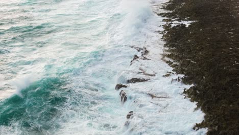 Bird's-eye-view-above-ocean-waves-crashing-on-steep-drop-off-section-misting-spray-over-rocks