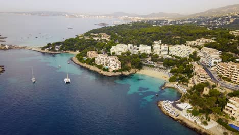 Playa-illetas-with-clear-turquoise-waters-and-anchored-yachts,-surrounded-by-greenery-and-resorts,-aerial-view