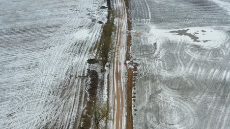 Aerial-follow-gravel-path-along-snow-covered-agriculture-grain-field-and-trees