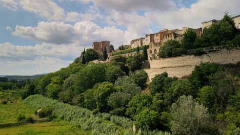 Drone-ascends-forested-hillside-above-farmland-fields-to-historic-town-archway-walls-of-Lauris-France