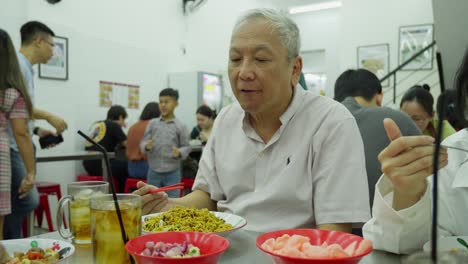 Un-Anciano-Asiático-Comiendo-Fideos-En-La-Mesa-Del-Restaurante-Con-Gente-Alrededor