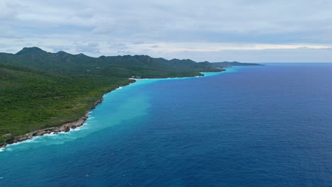 Lush-green-tropical-Caribbean-mountains-slope-to-grand-sea-cliffs-at-sunset,-panoramic-aerial-wide-angle-drone-dolly