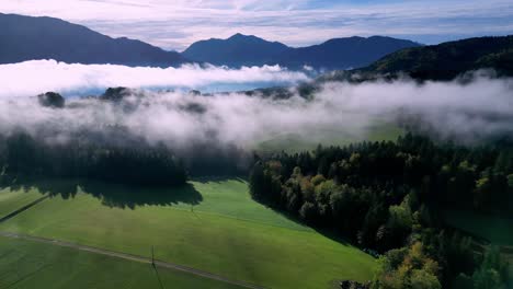 Aerial-view-of-a-lush-green-valley-filled-with-fog,-with-a-winding-lake-snaking-through-the-center-and-snow-capped-mountains-rising-in-the-distance