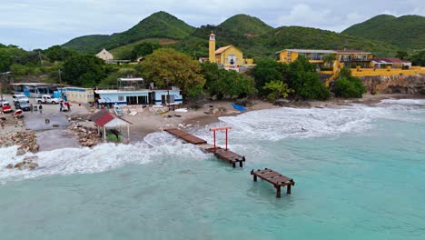 Wellen-Brechen-Auf-Einem-Kaputten-Holzsteg-Mit-Wanderhügeln-Und-Der-Ikonischen-Gelben-Küstenkirche-Auf-Der-Karibikinsel-Curaçao