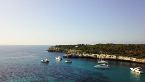 Approaching-drone-shot-moving-towards-the-picturesque-cove-where-sailboats,-and-yachts-are-docked-off-the-coast-of-Calla-Mondrago,-a-Natural-Park-located-in-Mallorca,-Spain