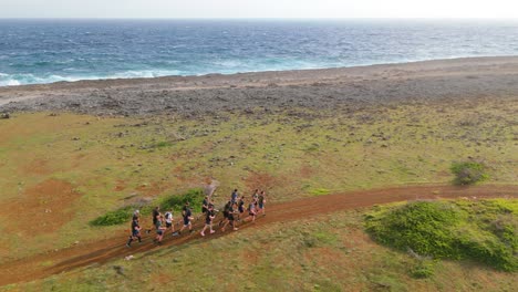 Panoramic-aerial-tracking-follows-large-group-of-men-training-and-running-on-dirt-road-along-empty-Caribbean-coastline