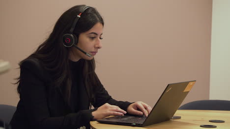 Businesswoman-in-meeting-room-using-headphones-to-talk-to-clients-while-looking-at-notes-on-her-laptop