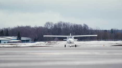 Small-white-ultralight-aircraft-taxiing-on-asphalt-apron-towards-camera-with-snow-in-background