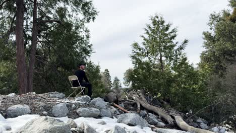 a-young-caucasian-male-dressed-in-black-hiking-clothes-and-a-black-ball-cap-walking-across-snow-and-rocks-in-the-forest-to-sit-on-a-plastic-chair-and-contemplate-life-on-an-overcast-day