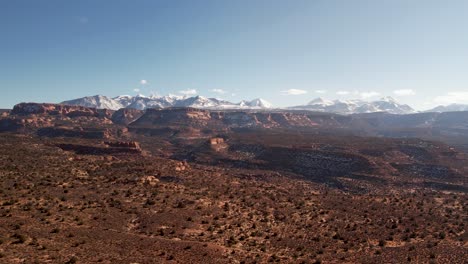 Eine-Hochfliegende-Drohne-Schoss-über-Eine-Abgelegene-Unbefestigte-Straße,-Die-Durch-Das-Weite-Und-Einzigartige-Wüstenland-In-Der-Nähe-Von-Moab,-Utah,-Führt,-Mit-Den-Schneebedeckten-Rocky-Mountains-In-Der-Ferne