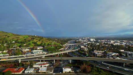 Rainbow-Skyline-above-slow-motion-highway-bridge-intersection-urban-traffic-city-of-clear-skyline,-aerial-drone-establishing-shot