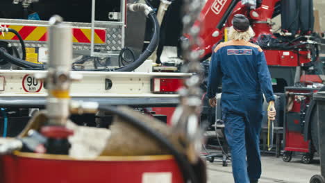 Female-car-mechanic-walking-towards-heavy-machinery-trucks-with-hoist-in-foreground