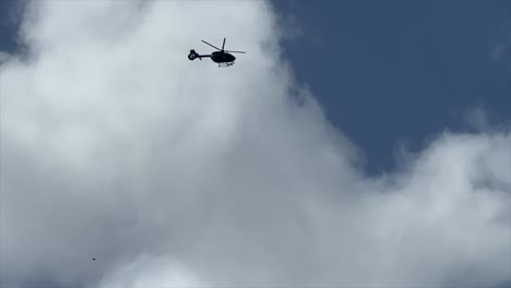 Slo-mo-of-Helicopter-flying-into-frame-from-top-left-overhead-with-puffy-clouds-behind-and-blue-sky-and-exits-frame-left