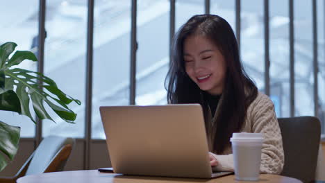 Joven-Asiática-De-Buen-Humor-Trabajando-En-Línea-En-Su-Computadora-Portátil-Desde-La-Cafetería