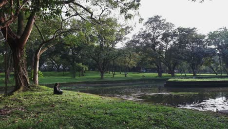 Solitary-girl-sitting-by-a-tranquil-lake-in-a-lush-park,-trees-reflecting-on-water-surface