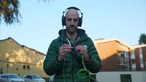 Bald-Male-Production-sound-mixer-putting-on-headphones-and-checking-levels-on-location-outside-in-urban-setting