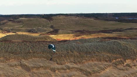 Toma-Aérea-Dinámica-Media-De-Un-Parapente-Navegando-Sobre-Dunas-De-Arena-Con-El-Duinreservaat-De-Noordhollands-Al-Fondo.