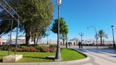 A-peaceful-park-in-Cádiz-features-well-manicured-gardens,-street-lamps,-and-pedestrians,-with-a-view-of-the-harbor-and-ships-in-the-distance