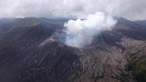 Elevándose-Por-Encima-De-Las-Nubes,-Revela-La-Impresionante-Actividad-Volcánica-Del-Monte-Bromo,-El-Humo-Ondulante-Y-Las-Erupciones-Ardientes-Crean-Una-Escena-Impresionante,-Imágenes-Aéreas-De-Drones-En-4k.