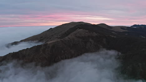 Picos-Montañosos-Brumosos-Al-Atardecer-Con-Cielos-Rosados-Suaves,-Vista-Aérea