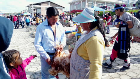 Un-Niño-Mira-Un-Pollo-Desplumado-En-Un-Mercado-En-Otavalo,-Ecuador,-Con-Gente-Alrededor,-Durante-El-Día.